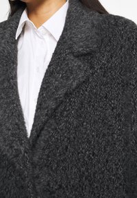 Cappotto nero e morbido con una finitura texturizzata e rever a forma di punta, indossato sopra una camicia bianca con bottoni. Inquadratura ravvicinata che mostra i dettagli del tessuto.