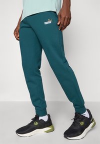 Pantalones jogger en color verde azulado, hechos de una tela suave, con bolsillos laterales y un pequeño logo de Puma en blanco y dorado en el muslo. Combinados con zapatillas deportivas negras.