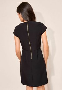 Fekete ruha rövid ujjakkal, testhez simuló felsőrész és bővülő szoknya. Jellemzője egy látható arany cipzár a hátsó részen. Simaságot sugalló szövettextúra.