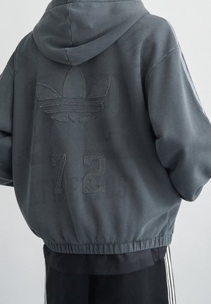 Personne portant un sweat à capuche gris foncé avec un grand logo cousu et le numéro 72 dans le dos, associé à un pantalon noir à rayures blanches.