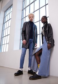 Jachetă de piele neagră și blugi pe o persoană; fustă din denim cu crăpătură înaltă și pantofi negri pe cealaltă. Lumină puternică dintr-o fereastră din spate.