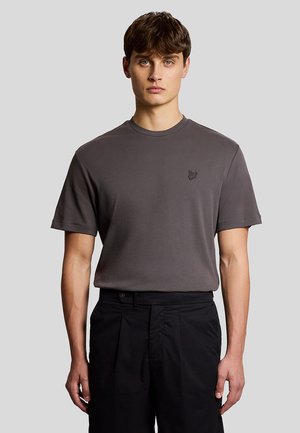 Ung mand iført en tætsiddende mørkegrå T-shirt med en lille broderet logo på brystet og sorte højttaljede bukser, stående mod en ensfarvet baggrund.