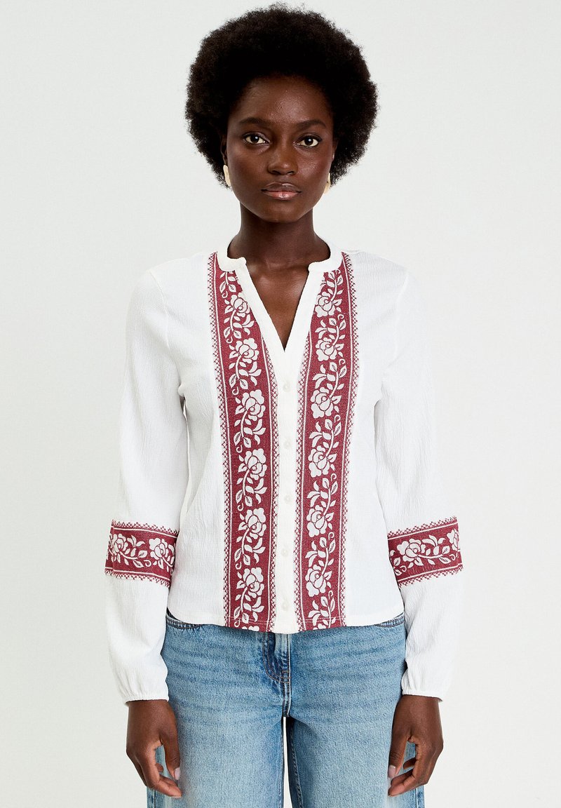 Blusa blanca con bordado floral rojo a lo largo del frente y las mangas, con escote en V y cierre de botones. Tela suave y con textura.