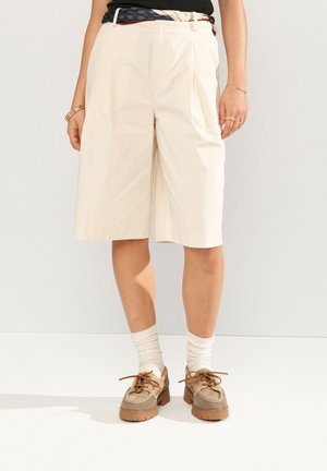 Person iført cremefarvede shorts til knæet, beige snøresko, hvide sokker og et mønstret bælte med smykker på synlige hænder.