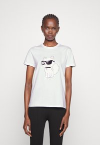T-shirt alb din bumbac cu imprimeu cu un lup desenat animat purtând ochelari de soare. Guler clasic rotund și mâneci scurte, asortat cu pantaloni negri cu dungi.