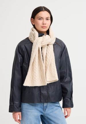 Sciarpa lavorata a maglia beige con un motivo a treccia, drappeggiata attorno al collo, abbinata a una giacca nera e jeans azzurri chiari. Aspetto tessurizzato e accogliente.