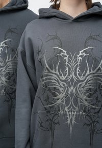 Sivá mikina s kapucňou z bavlneného zmesi, so strieborným grafickým dizajnom s prepracovanými vzormi na prednej strane a klokanie vrecko.