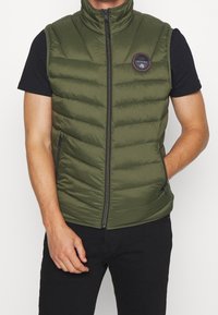 Olijfgroen gewatteerd mouwloos vest met een hoge kraag, ritsluiting aan de voorkant, ritszakken en een ronde logo patch op de borst.