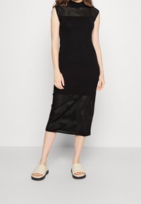 Robe noire longueur genou avec une texture côtelée et un col montant. Associée à des sandales slides beige. Présente une silhouette ajustée.