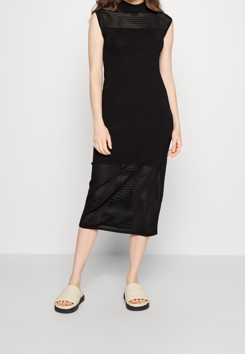 Robe noire longueur genou avec une texture côtelée et un col montant. Associée à des sandales slides beige. Présente une silhouette ajustée.