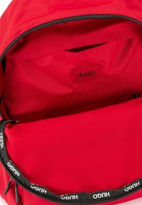 Interno di uno zaino rosso con un ampio scomparto, etichetta in tessuto con scritto "HUGO" e una cerniera nera con un logo bianco lungo il bordo.