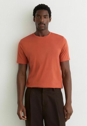 Mand står foran en hvid væg iført en orange t-shirt med korte ærmer og mørkebrune bukser, vender fremad med et neutralt udtryk.