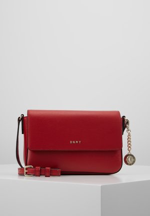 Sac bandoulière DKNY en cuir rouge avec sangle réglable et breloque chaîne dorée, présenté sur une surface blanche avec un fond gris.