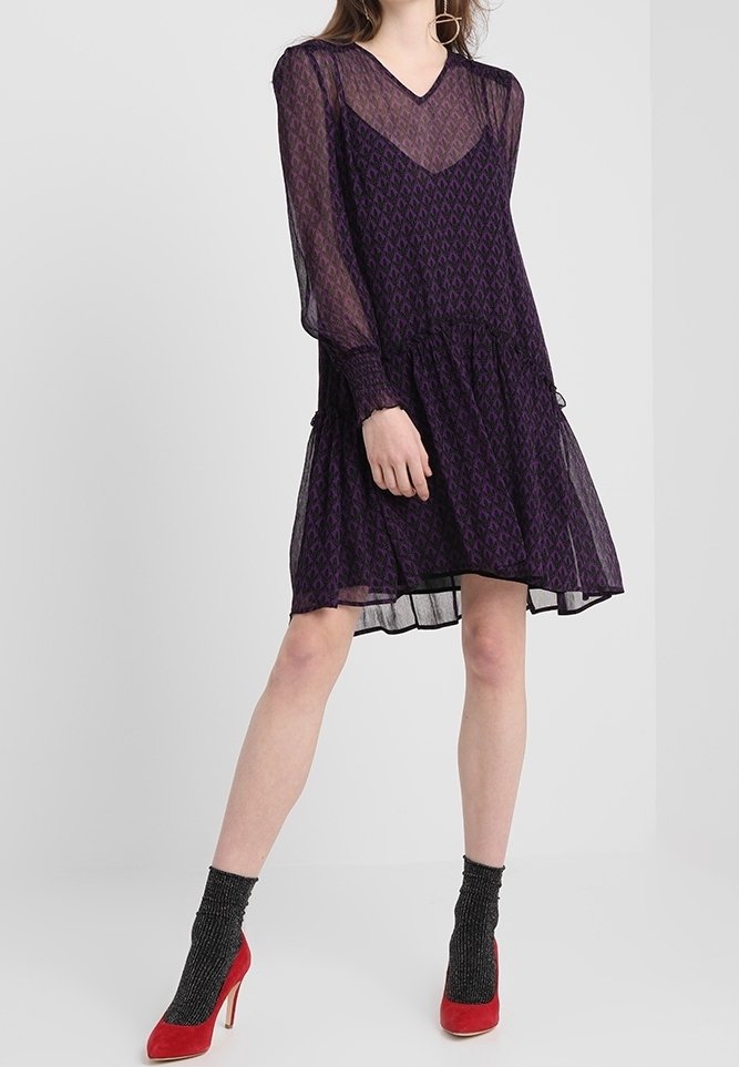 Robe violette transparente avec des manches longues, décolleté en V et jupe superposée arborant un motif en diamant. Associée à des escarpins rouges et des chaussettes noires texturées.