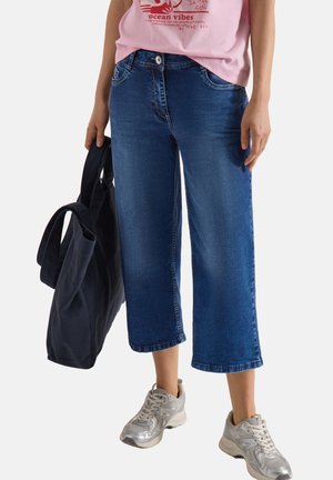 Persoon draagt cropped blauwe jeans, zilveren sneakers en een roze T-shirt met print, houdt een grote zwarte tas naast zich vast.