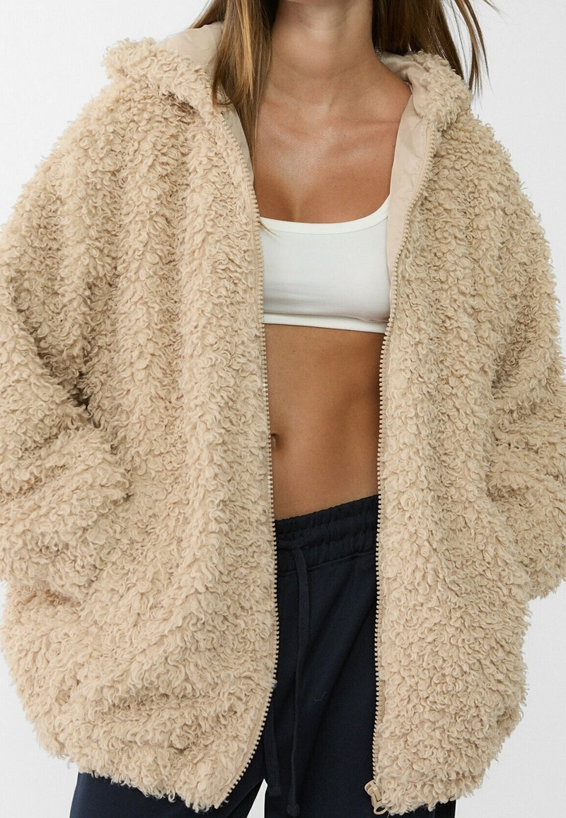 Beige, fluffy hooded jacket with textured fabric, featuring a full zipper and inseam pockets. Lined with smooth, matching beige material.