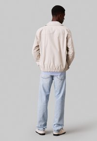 Giacca di cotone beige chiaro con colletto e orlo elastico; abbinata a jeans di denim blu chiaro e scarpe beige, vista dal retro.