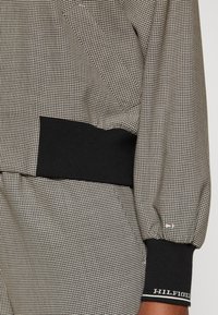 Außenbekleidung mit schwarz-weißer Karomusterung, schwarzem geripptem Bündchen und Saum, mit einem kleinen Logodetail am Ärmel.