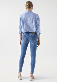 Sininen ja valkoinen raidallinen nappipaita, vaaleansiniset slim fit -farkut takataskuilla ja kreeminväriset korkeat sandaalit. Yksinkertainen valkoinen tausta.