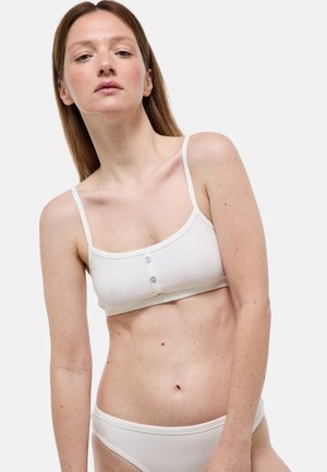 Femme portant un soutien-gorge côtelé blanc et une lingerie assortie sur un fond clair uni.