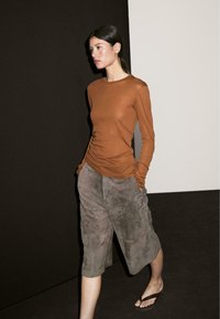 Woman with dark hair in a ponytail wears a brown long-sleeve top, grey knee-length loose pants, and brown flip-flops, walking indoors.