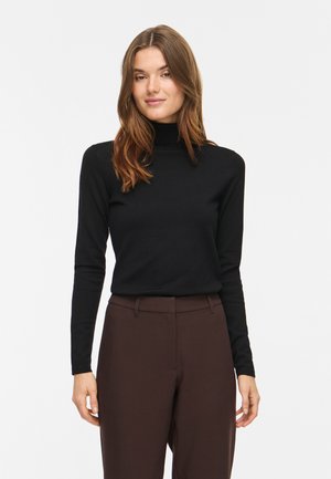 Femme aux longs cheveux bruns portant un pull à col roulé noir et un pantalon taille haute marron foncé, debout devant un fond blanc uni.