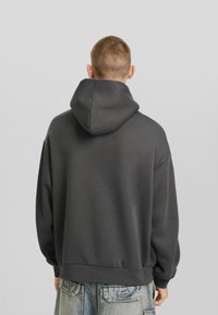 Felpa grigia oversize con cappuccio regolabile, realizzata in tessuto morbido e liscio, con polsini a costine. Il modello è di spalle e indossa jeans azzurri chiari.