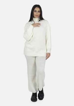 Suéter de punto grueso en color crema con cuello alto y textura acanalada, combinado con pantalones holgados a juego, complementados con botas negras.