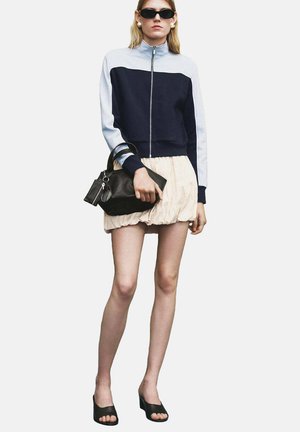 Navy and light blue zip-up jacket, cream skirt, black handbag, and black platform sandals. Model stands with arms at sides.