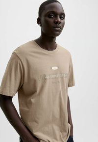 Mlada osoba s kratkom kosom koja nosi bež majicu s natpisima "1990" i "JACK & JONES", stojeći nasuprot jednostavne svijetle pozadine.