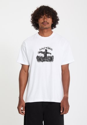 Homme aux cheveux bouclés portant un t-shirt blanc avec un motif d'épouvantail noir et le texte "Nineteen 90 Nowhere" et "Mandilux".