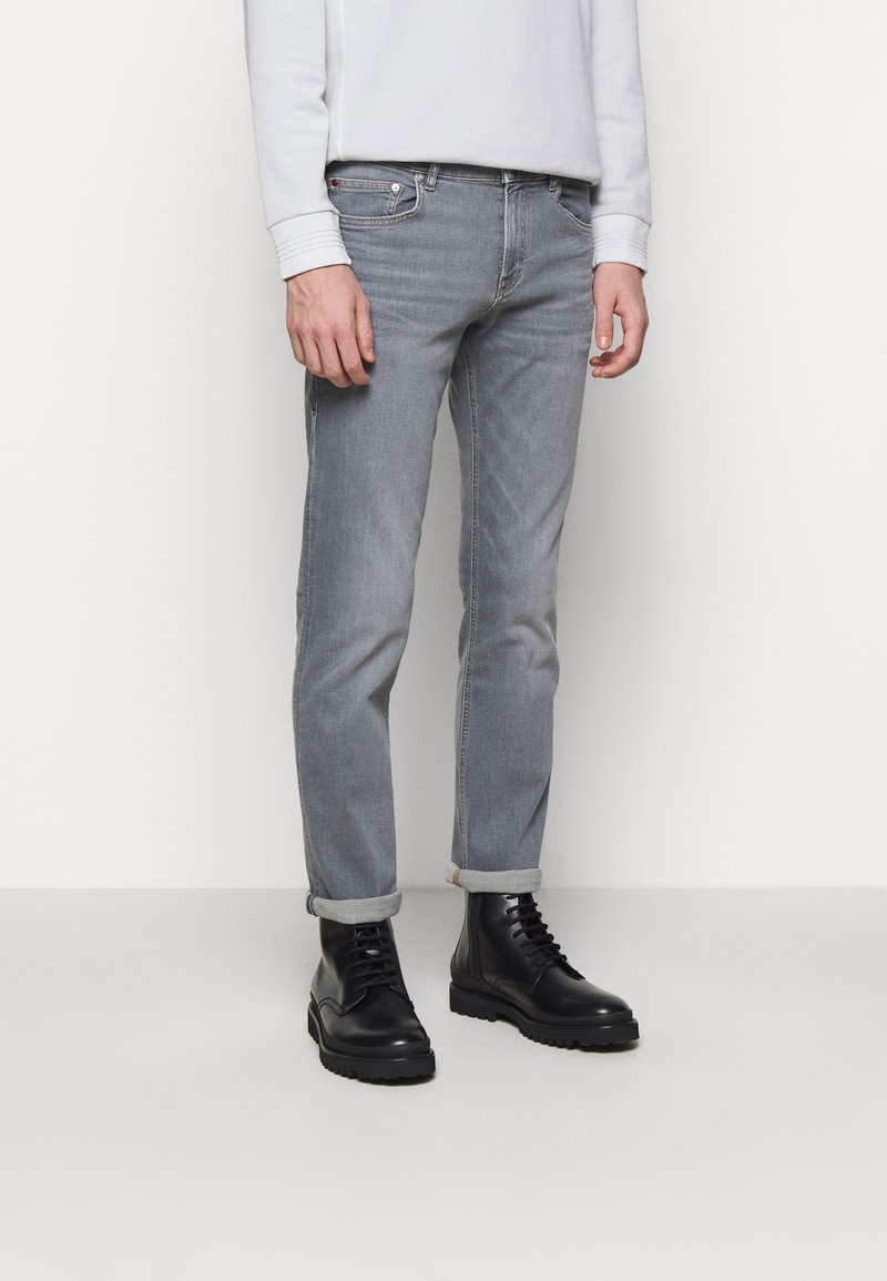 Homme portant un jean gris clair roulé et des bottes en cuir noir à lacets, debout devant un fond blanc uni.