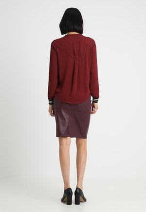 Kobieta stojąca tyłem, ubrana w bordową bluzkę z długim rękawem, ciemnofioletową skórzaną ołówkową spódnicę oraz czarne buty na słupku.