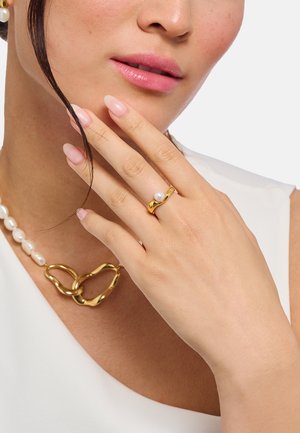 Woman with light pink nails wearing a gold ring with a pearl, and a white pearl and gold chain necklace, touching her face gently.