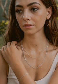 Silberne Halskette mit zwei Schichten, eine gebogen, die andere flach. Einfaches Silberarmband. Das Model trägt Creolen und ein leichtes Tops.
