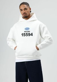 Witte hoodie met een voorzak, met een zwart gedrukt logo en de tekst "POLIZEI 15594". Gecombineerd met een donkere spijkerbroek.