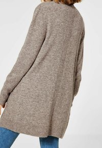 Žena oblečená v dlouhém, texturovaném taupe svetru s žebrovanými švy po zadní straně a na manžetách, v kombinaci se středně tmavými modrými džínami.