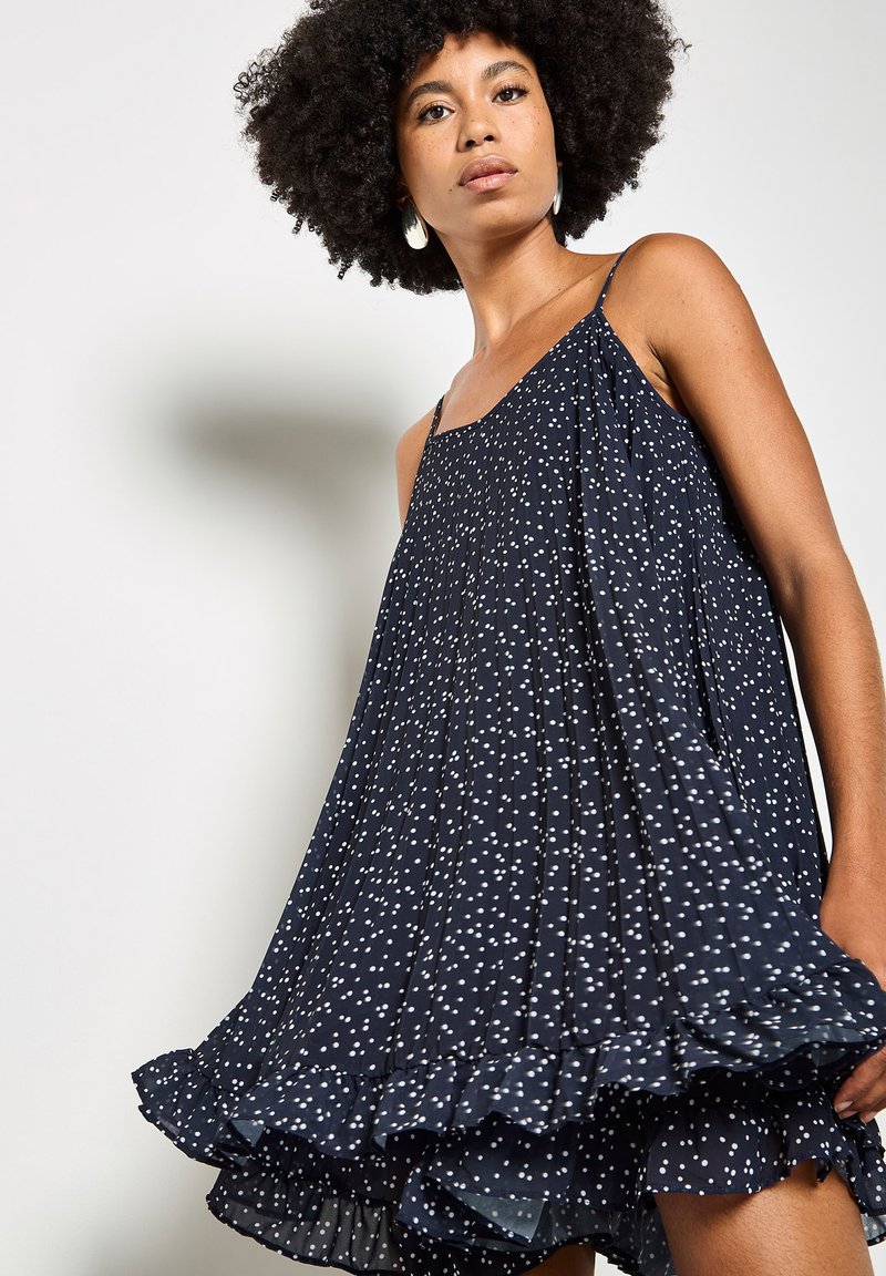 Femme aux cheveux bouclés portant une robe ample plissée bleu marine à pois blancs avec un ourlet volanté, debout devant un fond clair uni.