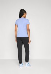 Kvinne står med ryggen til iført en lyseblå kortermet skjorte, svarte joggebukser, hvite sokker og hvite sneakers med blå detaljer.