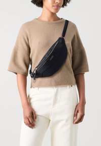 Schwarze Leder-Crossbody-Tasche mit strukturiertem Oberflächen, goldfarbener Reißverschluss, Logodetail und verstellbarem Schulterriemen, getragen über einem beigen Pullover und weißen Hosen.