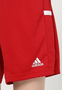 Bermudas deportivas rojas hechas de tejido liso con un panel de malla y el logo blanco de Adidas en la parte inferior izquierda. Cuentan con un bolsillo lateral con ribete blanco.