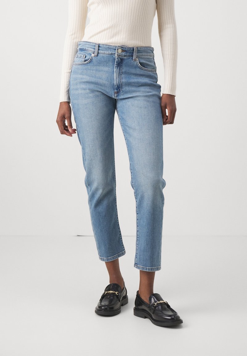 Jean droit taille haute bleu clair avec cinq poches et fermeture à boutons devant, associé à des mocassins noirs avec détail de chaîne dorée.