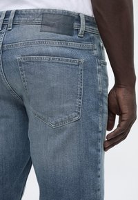 Πλάγια όψη ενός ατόμου που φοράει ανοιχτό μπλε τζιν παντελόνι με μαύρη ετικέτα Tom Tailor στη μέση και λευκό πουκάμισο μέσα από το παντελόνι.