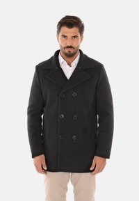 Cappotto peacoat nero in lana con chiusura a doppio petto, ampi rever, tasche laterali e vestibilità tailored. Indossato sopra una camicia bianca.