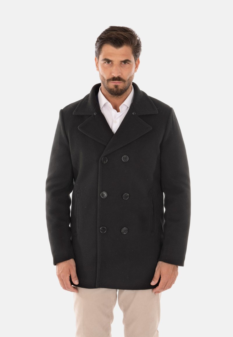 Cappotto peacoat nero in lana con chiusura a doppio petto, ampi rever, tasche laterali e vestibilità tailored. Indossato sopra una camicia bianca.