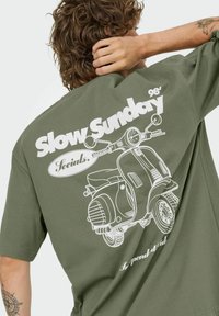 Olijfgroene t-shirt met een witte afbeelding van een vintage scooter en de tekst "Slow Sunday 98' Socials." Ronde halslijn en korte mouwen.
