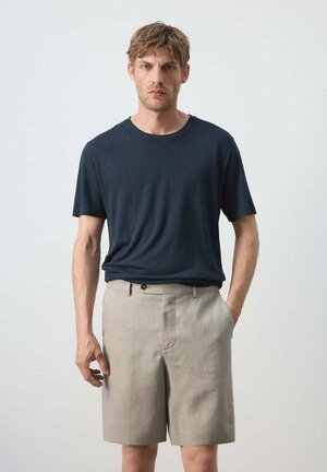 Homme portant une chemise bleu marine à manches courtes et un short beige à hauteur des genoux, debout avec une main dans la poche, sur un fond clair.