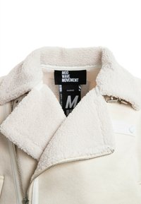 Chaqueta de piel sintética beige con un amplio cuello, cremallera plateada y etiqueta de logo. Textura suave y diseño resistente, adecuada para superponer.