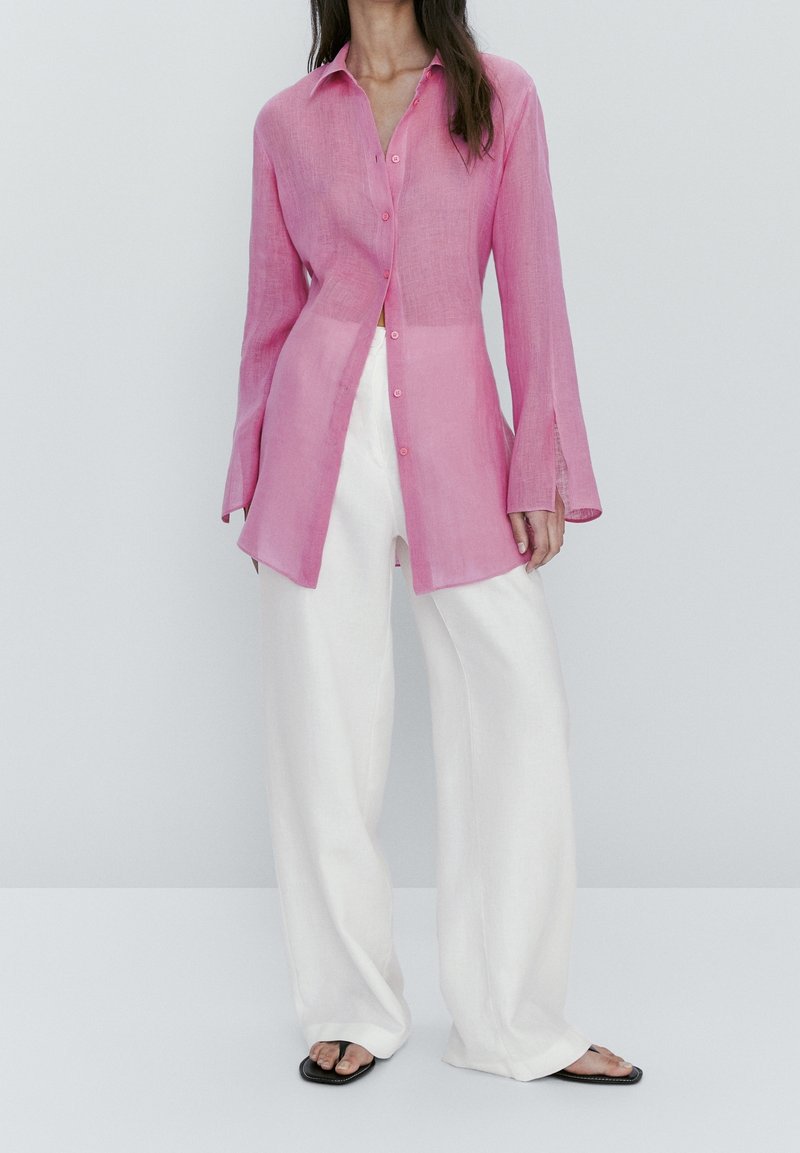 Femme portant une chemise rose ample boutonnée à manches longues, un pantalon blanc à jambes larges et des sandales noires à bout ouvert, debout devant un fond gris.