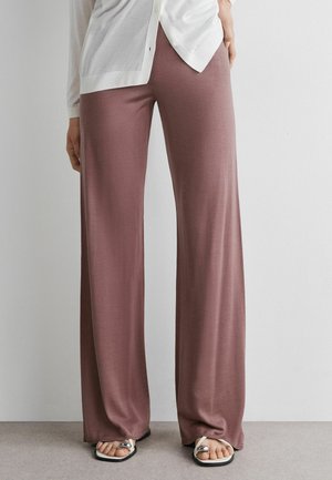 Personne portant un pantalon mauve à jambes larges, un cardigan blanc boutonné et des sandales blanches avec des accents argentés, debout contre un mur uni.